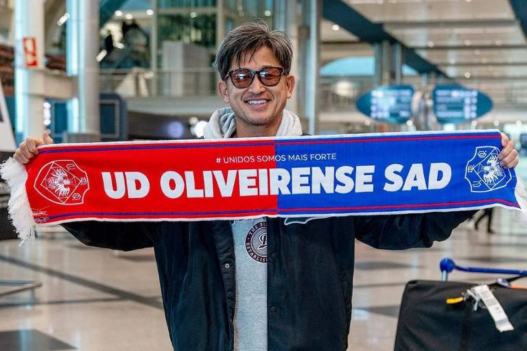 O atacante Kazu Miura, usando óculos e sorrindo, segura flâmula nas cores vermelha e azul com o nome do Oliveirense, clube de Portugal que ele defendeu em 2023 e 2024