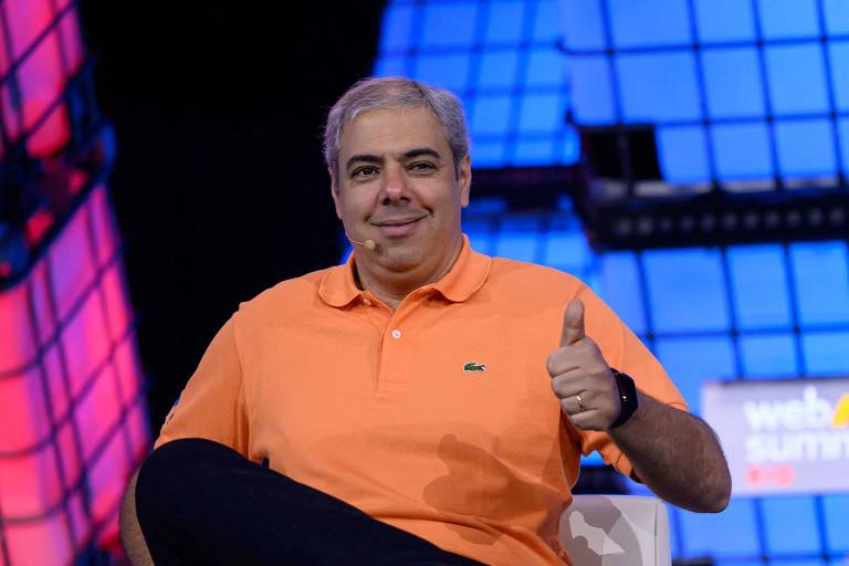 Um homem está sentado em uma cadeira branca, vestindo uma camisa polo laranja e calça preta. Ele está fazendo um gesto de positivo com a mão direita. O fundo é composto por luzes coloridas em tons de azul e rosa, com um painel que exibe o texto 'web summit'.