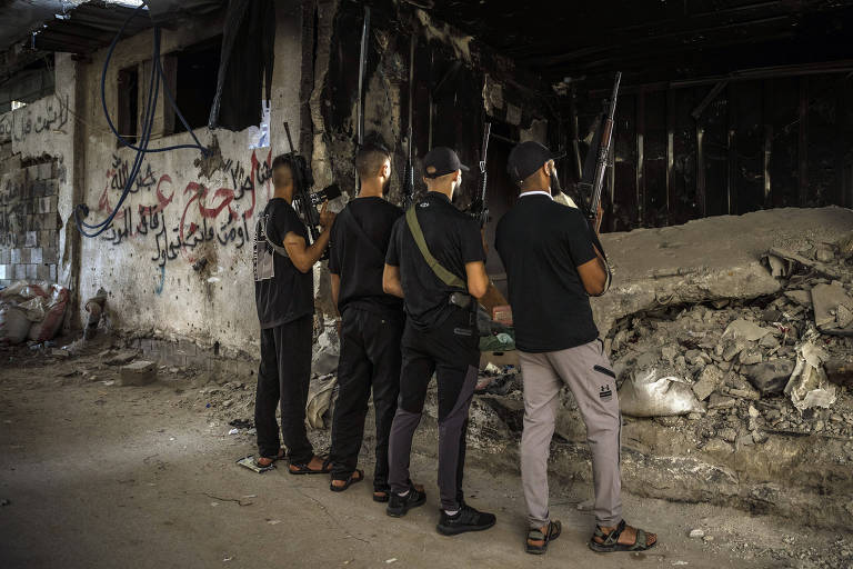 Quatro homens armados, vestidos de preto, estão de pé em frente a um prédio destruído. O prédio tem paredes danificadas e escombros no chão. Há grafites em árabe nas paredes do prédio.