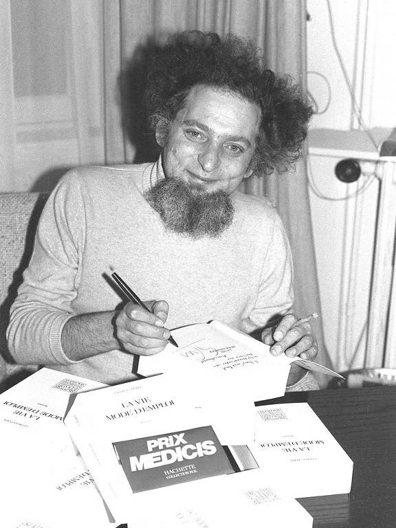 Fotografia em preto e branco de um homem com barba e cabelo volumoso, sentado à mesa, assinando livros. Ele está sorrindo e segurando uma caneta na mão direita. Na mesa, há vários livros empilhados
