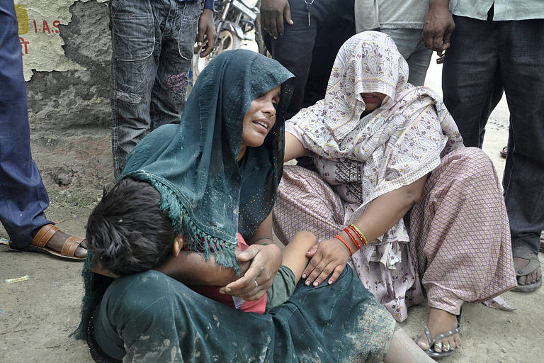 A imagem mostra duas mulheres sentadas no chão, visivelmente tristes. Uma delas está vestida com um sari verde e segura uma criança em seu colo. A outra mulher está coberta com um tecido estampado e também parece estar chorando. Há várias pessoas em pé ao redor delas.