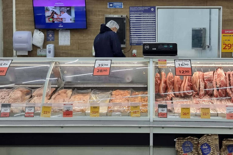 Imagem de um balcão de carnes em um supermercado. Há várias bandejas de carne expostas, com etiquetas de preço em vermelho. Acima do balcão, há uma série de placas com os preços de diferentes cortes de carne. Ao fundo, há um funcionário de avental branco e boné azul, trabalhando atrás do balcão. À esquerda, há uma máquina de cortar carne e uma tela de computador. À direita, há uma tela azul com informações e uma prateleira com produtos embalados.