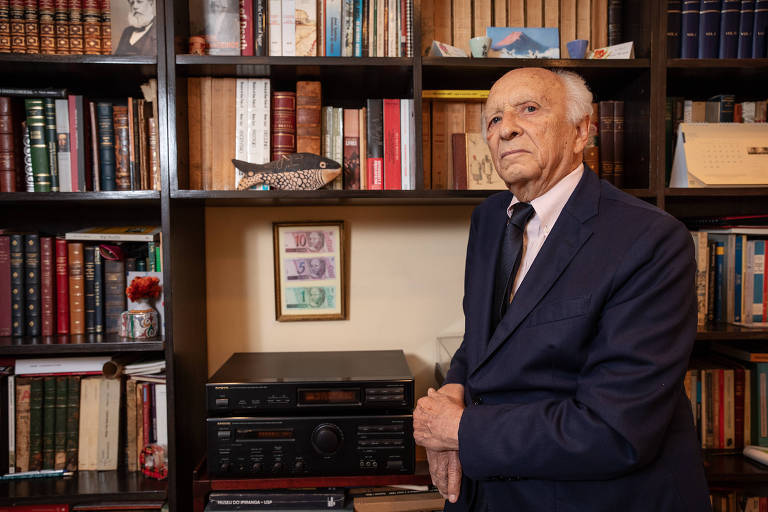 Homem idoso, de cabelos brancos, um pouco calvo, usando terno escuro, está de pé apoiado em uma estante de livros, cercado por livros