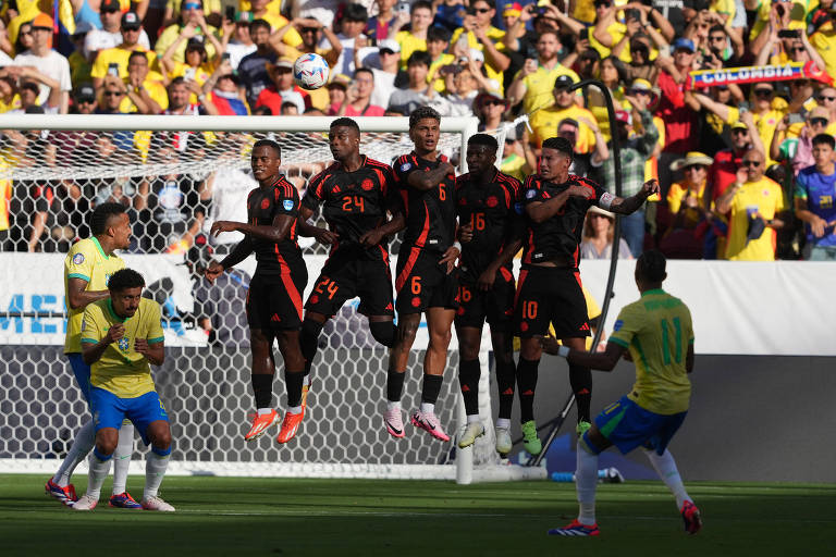 Jogador com camisa amarela e calção azul observa a bola passar sob uma barreira de cinco jogadores com o uniforme da Colômbia, preto, com detalhes em vermelho