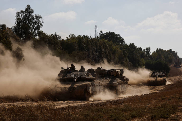 A imagem mostra três tanques militares em movimento em uma estrada de terra, levantando poeira enquanto avançam. Os tanques estão em uma área arborizada, com árvores ao fundo e um céu parcialmente nublado acima.