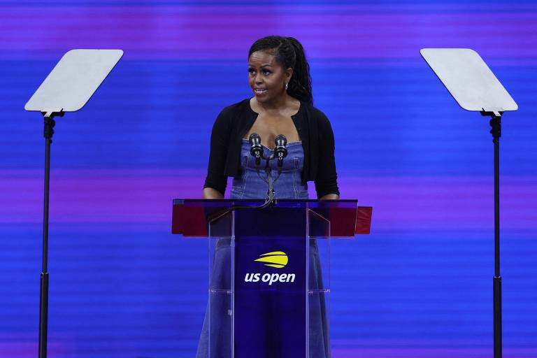 Uma mulher está discursando em um pódio com o logotipo do US Open. Ela está usando um vestido azul e um casaco preto. Há dois microfones no pódio e dois teleprompters transparentes posicionados à sua frente. O fundo é roxo.