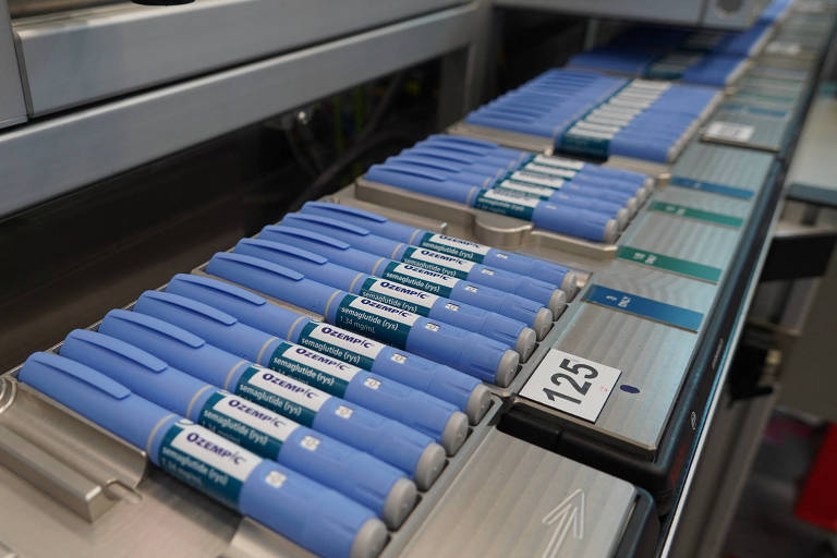 A imagem mostra uma linha de produção com várias canetas de insulina azuis organizadas em fileiras. As canetas estão alinhadas em uma esteira transportadora, prontas para serem embaladas ou distribuídas. Há etiquetas e números visíveis, como '125', indicando a organização do processo.
