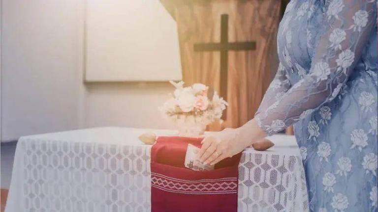 A imagem mostra uma pessoa arrumando um altar em uma igreja. A pessoa está vestindo uma roupa azul com detalhes florais e está ajustando um tecido vermelho sobre uma toalha branca que cobre o altar. Ao fundo, há uma cruz de madeira e um arranjo de flores sobre o altar.
