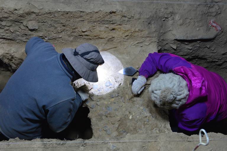 A imagem mostra dois arqueólogos trabalhando em uma escavação. Eles estão em um buraco, usando ferramentas para escavar cuidadosamente o solo. Um dos arqueólogos está vestindo um casaco azul e um chapéu cinza, enquanto o outro está usando um casaco roxo e um gorro cinza. 
