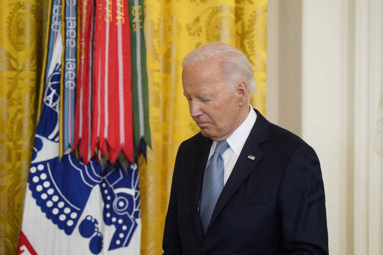 Biden, de terno escuro e gravata azul clara, está de pé em frente a um conjunto de bandeiras coloridas. Ele está olhando para baixo e parece estar em um ambiente formal, possivelmente uma cerimônia ou evento oficial. O fundo é decorado com uma cortina amarela com padrões.