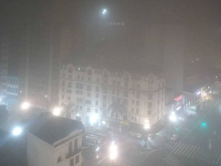 A imagem mostra uma cidade à noite coberta por uma densa neblina. As luzes dos postes e dos edifícios estão acesas, criando um efeito difuso devido à neblina. Vários edifícios são visíveis, mas suas formas estão parcialmente obscurecidas pela neblina. Há também algumas luzes de veículos nas ruas, indicando tráfego noturno.