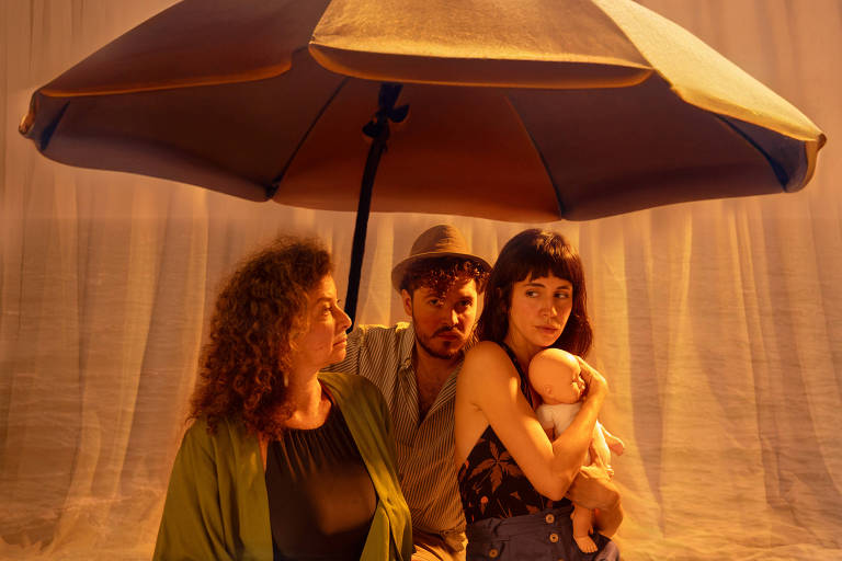 A imagem mostra três pessoas sentadas sob um grande guarda-sol. À esquerda, uma mulher de cabelos cacheados e vestindo uma blusa preta com um casaco verde. No centro, um homem de barba e cabelo cacheado, usando um chapéu e uma camisa listrada. À direita, uma mulher de cabelos escuros e franja, vestindo uma blusa estampada e segurando um bebê de brinquedo. O fundo é iluminado com uma luz quente, criando uma atmosfera acolhedora