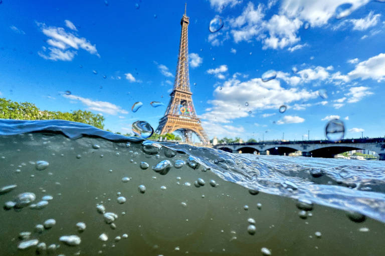A imagem mostra uma vista subaquática da Torre Eiffel em Paris, França. A foto é tirada parcialmente debaixo d'água, com bolhas visíveis na parte submersa. Acima da linha d'água, a Torre Eiffel se ergue contra um céu azul com algumas nuvens brancas. À direita, há uma ponte que atravessa o rio Sena.