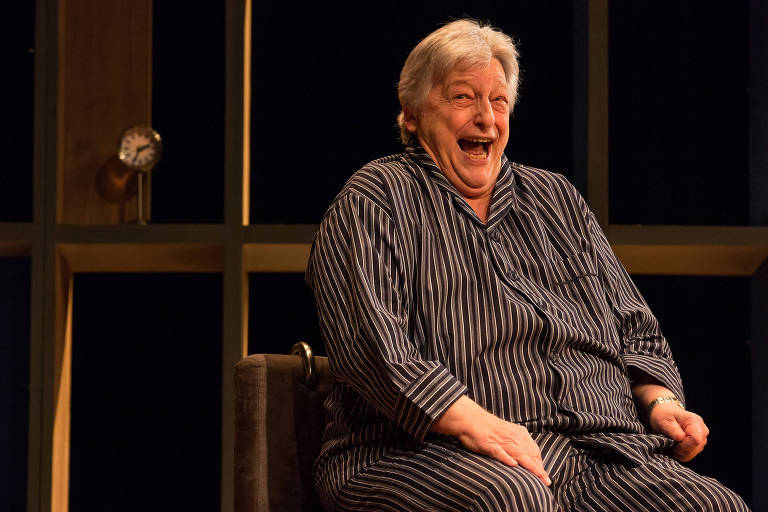homem branco idoso, usando pijama listrado, sentado em uma cadeira e rindo