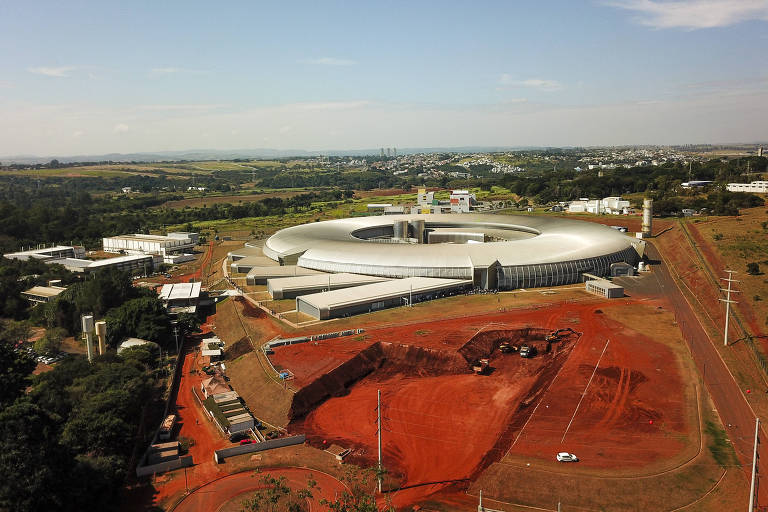 Imagem aérea de um complexo industrial com um grande edifício circular no centro. O solo ao redor do edifício é de terra vermelha, e há várias construções menores ao redor. A paisagem ao fundo é composta de áreas verdes e algumas construções ao longe.