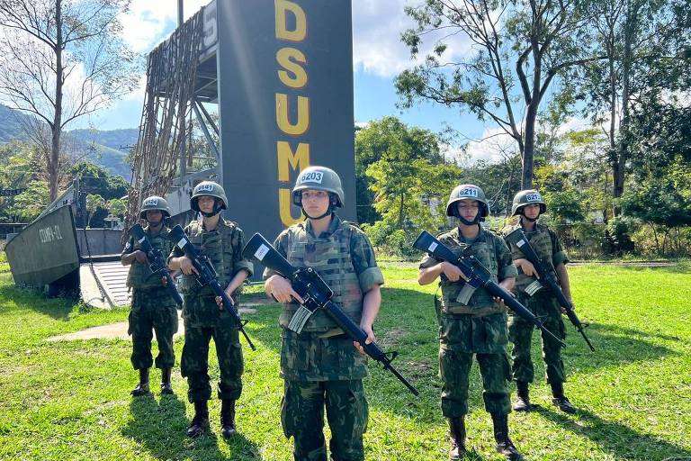 A imagem mostra cinco mulheres em uniforme militar, equipadas com capacetes e armas, posando em frente a uma estrutura vertical com a inscrição 'DSUM'. Elas estão em um campo gramado com árvores ao fundo e montanhas ao longe.