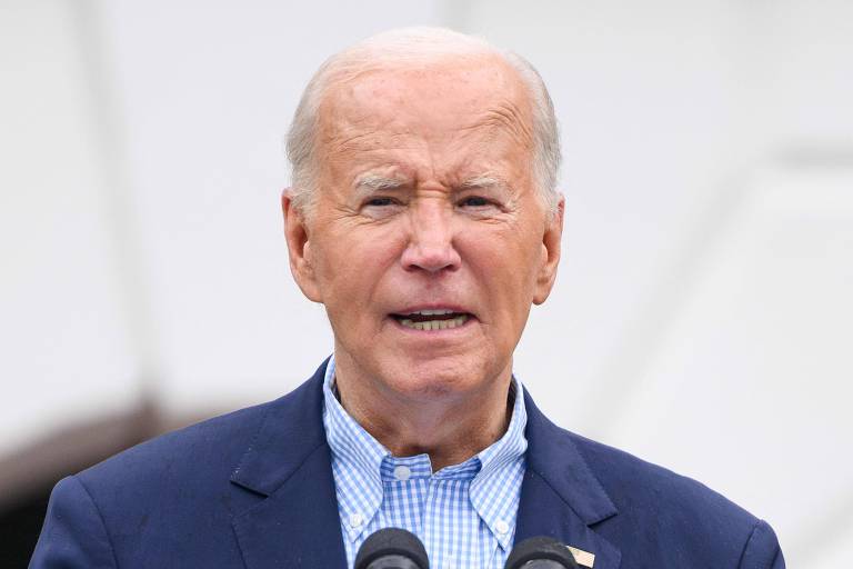 A imagem mostra Biden, um homem branco idoso de cabelo branco e curto, vestindo um terno azul escuro e uma camisa azul clara com padrão xadrez. Ele está falando ao microfone e está ao ar livre, com um fundo desfocado.
