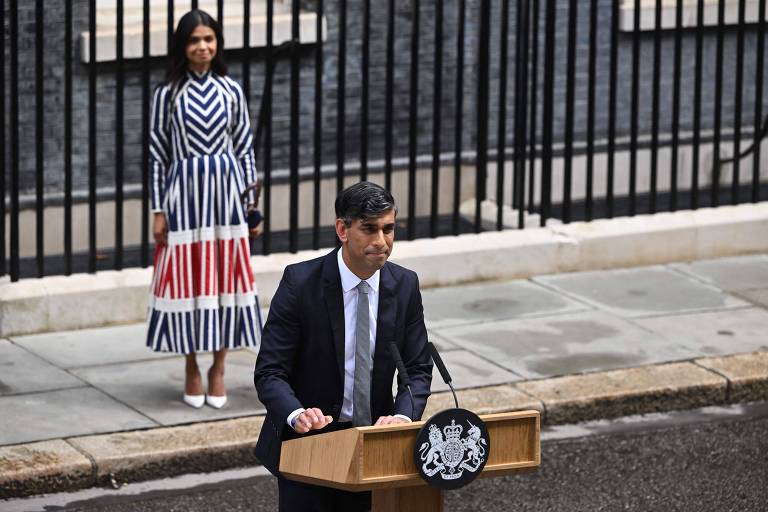 Um homem de terno escuro e gravata está fazendo um discurso em um púlpito com o emblema do governo britânico. Atrás dele, uma mulher está de pé, vestindo um vestido longo com listras verticais em azul, branco e vermelho. Eles estão em frente a um prédio com grades pretas.