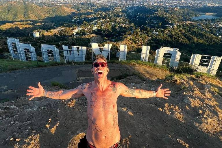 Um homem sem camisa, usando óculos de sol, está de braços abertos e sorrindo em frente ao famoso letreiro de Hollywood, localizado em Los Angeles, Califórnia. A imagem é tirada de um ponto elevado, mostrando uma vista panorâmica da cidade ao fundo, com colinas verdes e áreas urbanas ao longe.