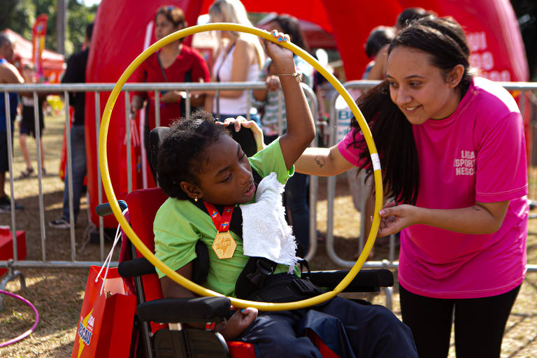 Uma menina negra, cadeirante brinca com um bambolê amarelo; ela é auxiliada por uma jovem que usa camiseta rosa; ao fundo, se vê outras pessoas transitando