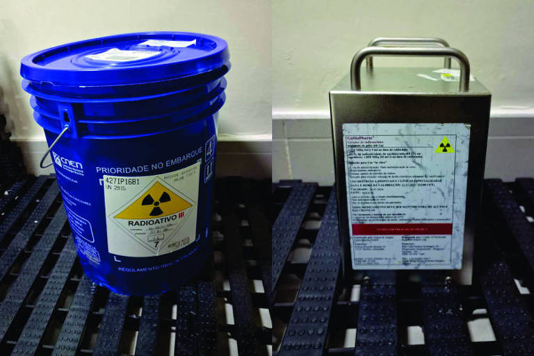 A imagem mostra três recipientes diferentes, todos com etiquetas de aviso de material radioativo. À esquerda, há um balde azul com uma tampa, no centro, um recipiente metálico com uma alça e, à direita, um balde branco com uma tampa. Todos os recipientes estão sobre uma superfície gradeada.