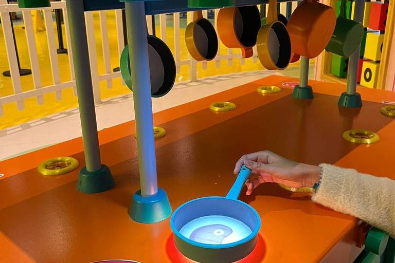 Imagem de um brinquedo de cozinha infantil com uma bancada laranja e utensílios pendurados. Uma mão segura uma frigideira azul sobre uma luz circular na bancada. Há um adesivo de melancia na bancada e botões coloridos ao redor. Ao fundo, há uma cerca branca e um ambiente com piso amarelo.
