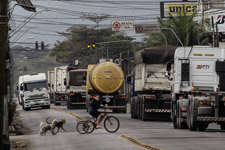 A imagem mostra uma estrada com um intenso tráfego de caminhões. No centro da imagem, um ciclista atravessa a rua com dois cães. Há um caminhão-tanque com a imagem de uma laranja na traseira. Placas de trânsito e fios elétricos são visíveis ao longo da estrada. No fundo, há uma placa com o texto 'unic' e outra com o texto 'NESTA VIA'.
