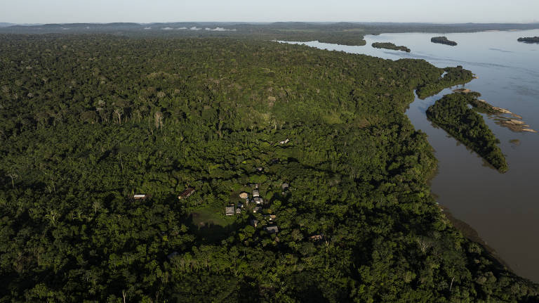 Imagem aérea de uma vasta área de floresta amazônica com uma pequena aldeia no centro. A aldeia é composta por várias construções, incluindo casas e outras estruturas, cercadas por vegetação densa. À direita da imagem, há um grande rio com várias ilhas cobertas de vegetação. O horizonte mostra uma extensão contínua de floresta.