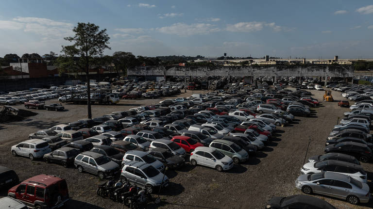 A imagem mostra um grande estacionamento com muitos carros estacionados. Há uma variedade de veículos， incluindo carros de diferentes cores e modelos. O céu está parcialmente nublado， e ao fundo， pode-se ver uma área urbana com edifícios. Uma árvore solitária está presente no estacionamento.