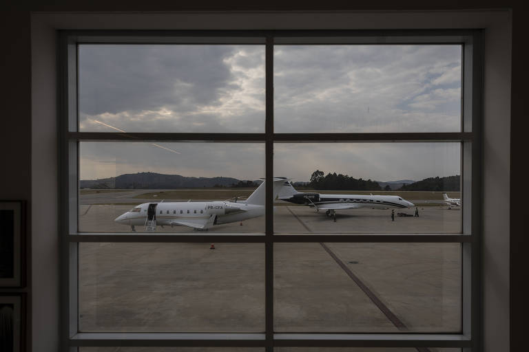A imagem mostra uma vista de um aeroporto através de uma janela com divisórias. No pátio, há dois aviões estacionados. O céu está nublado e ao fundo é possível ver colinas e vegetação.