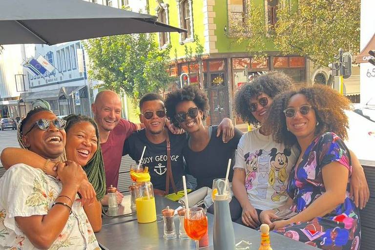 Um grupo de sete pessoas está sentado em uma mesa ao ar livre em um café. Todos estão sorrindo e parecem estar se divertindo. Há várias bebidas na mesa, incluindo coquetéis e sucos. Ao fundo, há um edifício verde com arquitetura clássica e outras construções urbanas. A mesa está coberta por um guarda-sol e há árvores ao redor.