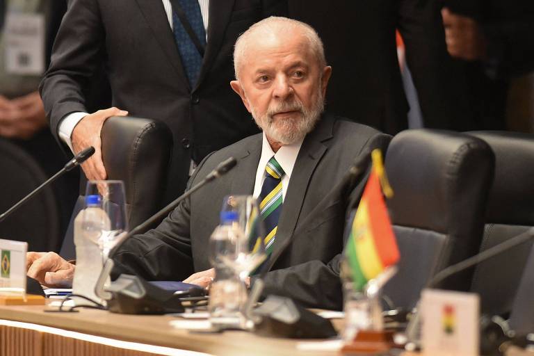 Lula, um homem idoso com barba branca, está sentado em uma mesa de conferência. Ele veste um terno escuro com uma gravata listrada em verde e amarelo. Na mesa, há garrafas de água, copos e bandeiras de diferentes países. Ao fundo, outras pessoas estão presentes, mas desfocadas.