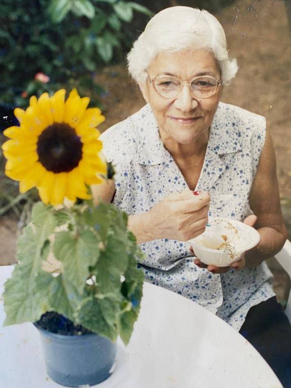 A imagem mostra uma senhora idosa de cabelos brancos e óculos, sentada ao ar livre. Ela está segurando uma tigela e uma colher, parecendo estar comendo algo. Em primeiro plano, há um girassol em um vaso sobre a mesa. Ao fundo, há vegetação verde.
