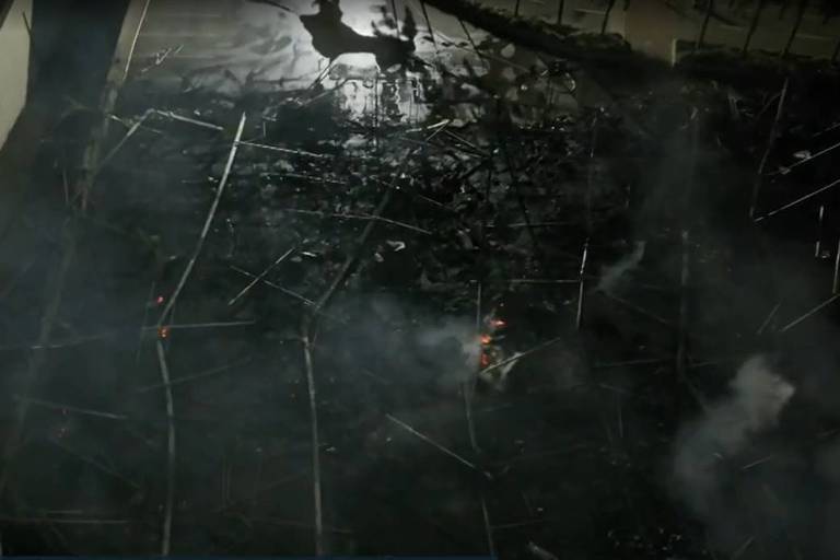 Imagem aérea mostra os destroços de uma estrutura queimada, com restos de metal e madeira espalhados pelo chão. Há fumaça e pequenos focos de fogo ainda visíveis. A imagem é de uma transmissão ao vivo de um noticiário.