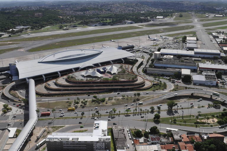 Imagem aérea do aeroporto de Recife com várias pistas de pouso e decolagem visíveis. O terminal principal tem um design moderno e está cercado por estradas e estacionamentos. Há várias construções ao redor do terminal, e a paisagem ao fundo é composta por áreas verdes e algumas construções.