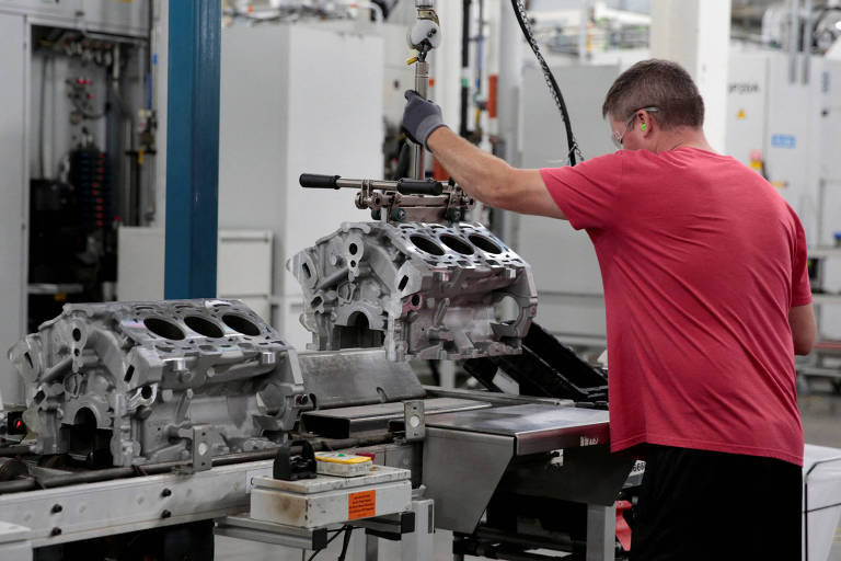 Um trabalhador, vestindo uma camiseta vermelha e luvas, está operando uma máquina em uma linha de montagem de motores em uma fábrica. Ele está manipulando uma ferramenta sobre um bloco de motor, que está posicionado em uma esteira. O ambiente ao redor é industrial, com várias máquinas e equipamentos visíveis.