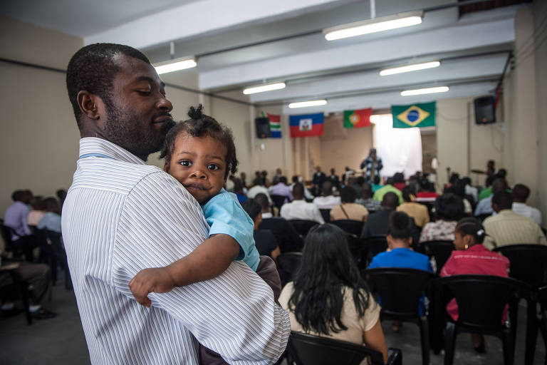Um homem segura um bebê em um evento comunitário realizado em um salão. Ao fundo, várias pessoas estão sentadas em cadeiras de plástico preto, assistindo a uma apresentação ou reunião. Na parede ao fundo, há três bandeiras penduradas: a bandeira do Haiti, a bandeira de Portugal e a bandeira do Brasil.