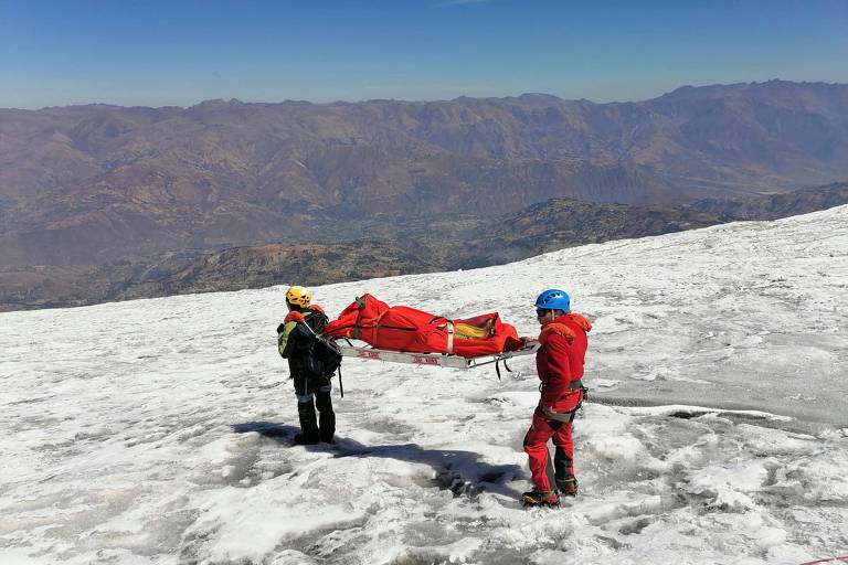Dois socorristas carregam uma maca com o corpo de um alpinista, coberto por uma lona laranja, em uma montanha cujo solo é coberto de neve. Ao fundo, é possível ver mais montanhas, sem neve, e o céu azul