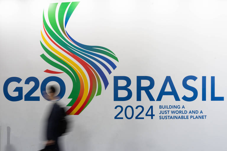 Imagem mostra um homem passando em frente a um mural onde está a logomarca do G20 Brasil, com listras sinuosas verticalmente de várias cores