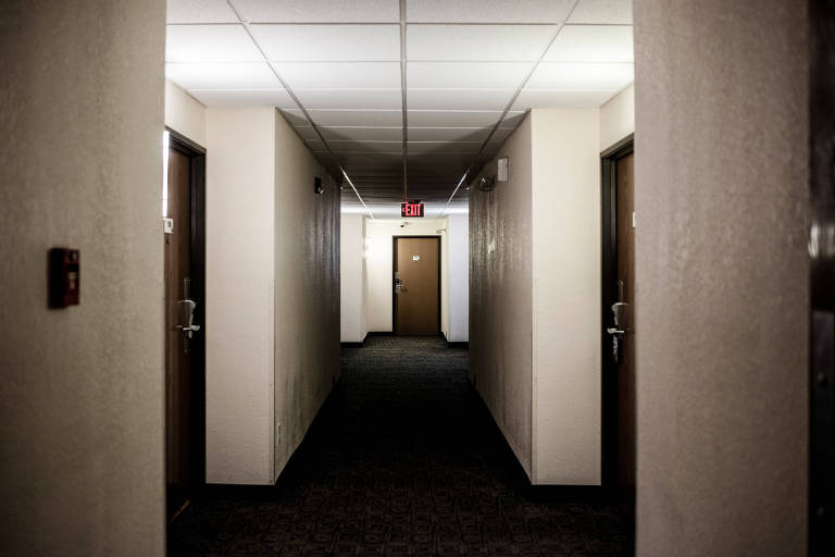 A imagem mostra um corredor de hotel com paredes claras e piso escuro. Há várias portas de madeira ao longo do corredor, todas fechadas. No final do corredor, há uma porta adicional e uma luz de saída vermelha no teto.