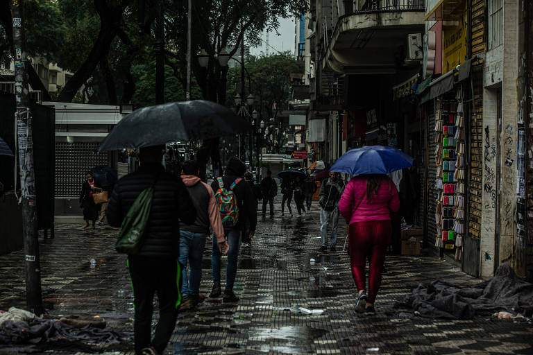 A imagem mostra uma rua movimentada em um dia chuvoso. Várias pessoas estão caminhando com guarda-chuvas abertos. O chão está molhado e reflete a luz ambiente. À direita, há lojas com vitrines e letreiros. Árvores estão presentes ao fundo, e o céu está nublado.