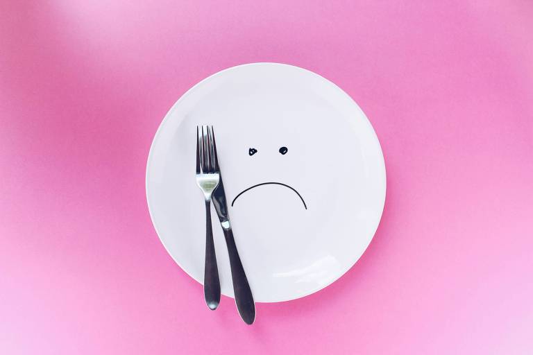 Fotografia colorida mostra prato branco com rosto triste desenhado ao centro. Sobre um fundo cor-de-rosa há um garfo e uma faca