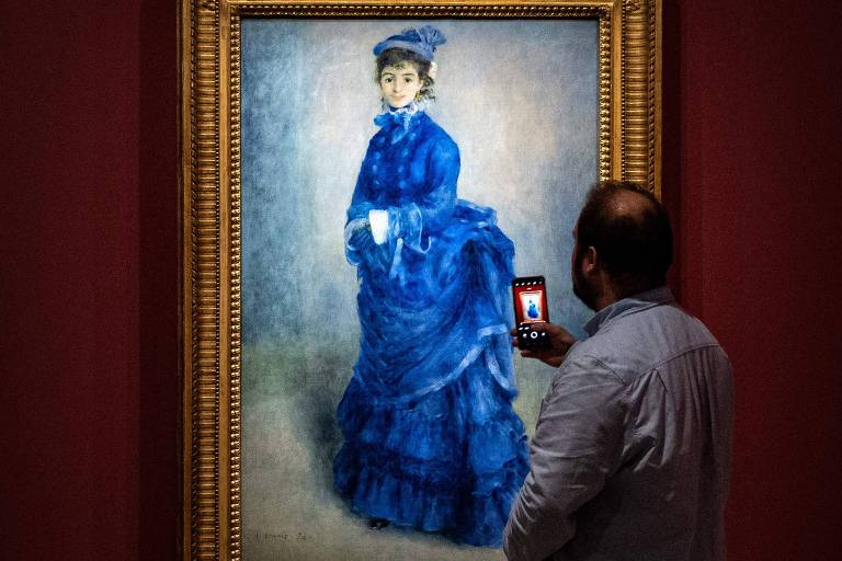A imagem mostra um homem de pé em frente a uma pintura emoldurada em um museu. Ele está tirando uma foto da pintura com seu smartphone. A pintura retrata uma mulher vestida com um vestido azul e um chapéu combinando, em um fundo neutro. A moldura da pintura é dourada e a parede do museu é de cor vermelha escura.