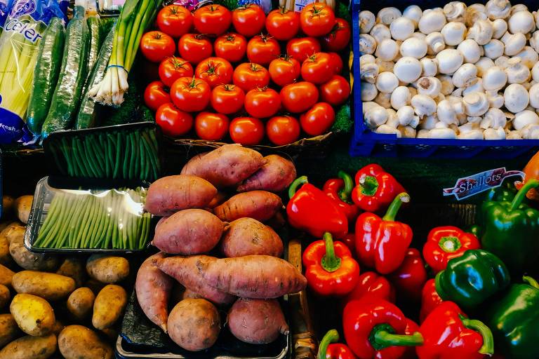 Imagem de uma banca de vegetais em um mercado. Há uma variedade de vegetais, incluindo tomates, cogumelos, pimentões vermelhos e verdes, batatas, batatas-doces, feijões verdes, cebolinhas, pepinos embalados e outros vegetais embalados.