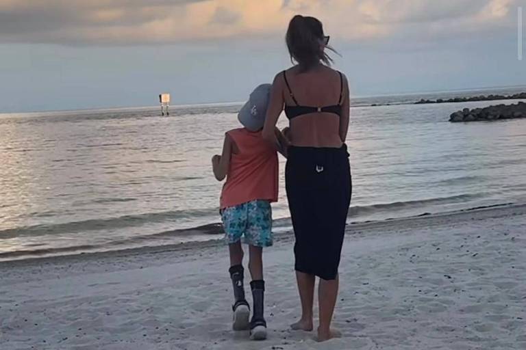 Mulher de biquíni caminha com menino com necessidades especiais na areia de uma praia