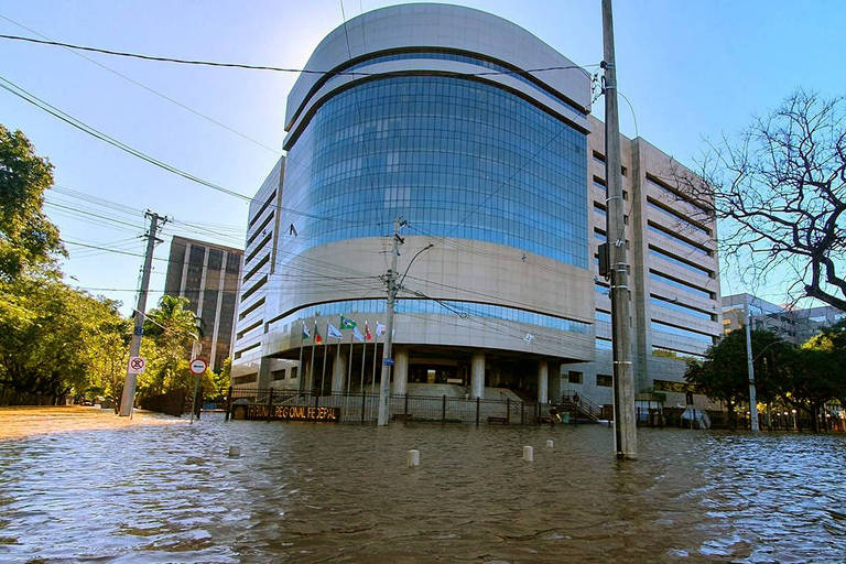 Na foto, um prédio de esquina com detalhes espelhados está ilhado. A água da enchente circunda toda a área externa