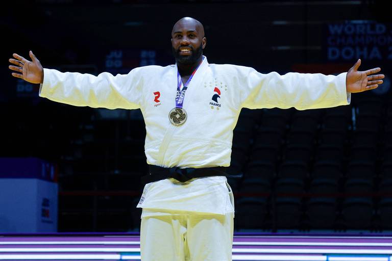 Um judoca está de pé com os braços abertos, vestindo um quimono branco com uma faixa preta. Ele está usando uma medalha ao redor do pescoço. No quimono, há um logotipo vermelho e a palavra 'France' com um símbolo. Ao fundo, há uma arquibancada vazia e um banner com as palavras 'World Champion Doha'.