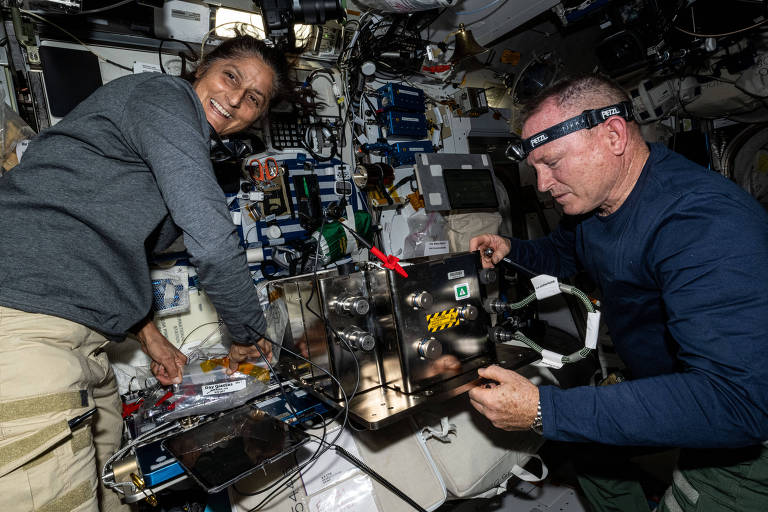 Dois astronautas estão trabalhando dentro da Estação Espacial Internacional. Um astronauta está segurando um dispositivo, enquanto o outro está sorrindo e ajudando. O ambiente é cheio de equipamentos científicos e painéis de controle
