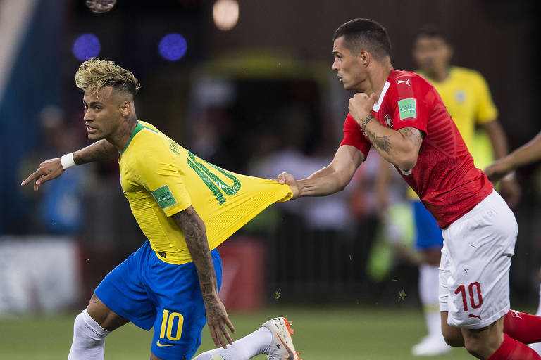 Imagem de um jogo de futebol onde o jogador Neymar, com a camisa amarela e o número 10, está sendo puxado pela camisa por um jogador adversário com a camisa vermelha e o número 10. Neymar usa um grande topete loiro, com a lateral da cabeça raspada