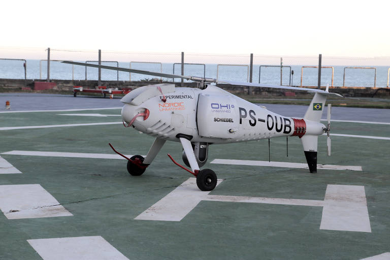 A imagem mostra um drone de grande porte estacionado em um heliponto. O drone é branco com detalhes em vermelho e preto, e possui a inscrição 'PS-DUB' em sua lateral. Ao fundo, é possível ver uma cerca e o horizonte com o céu claro.